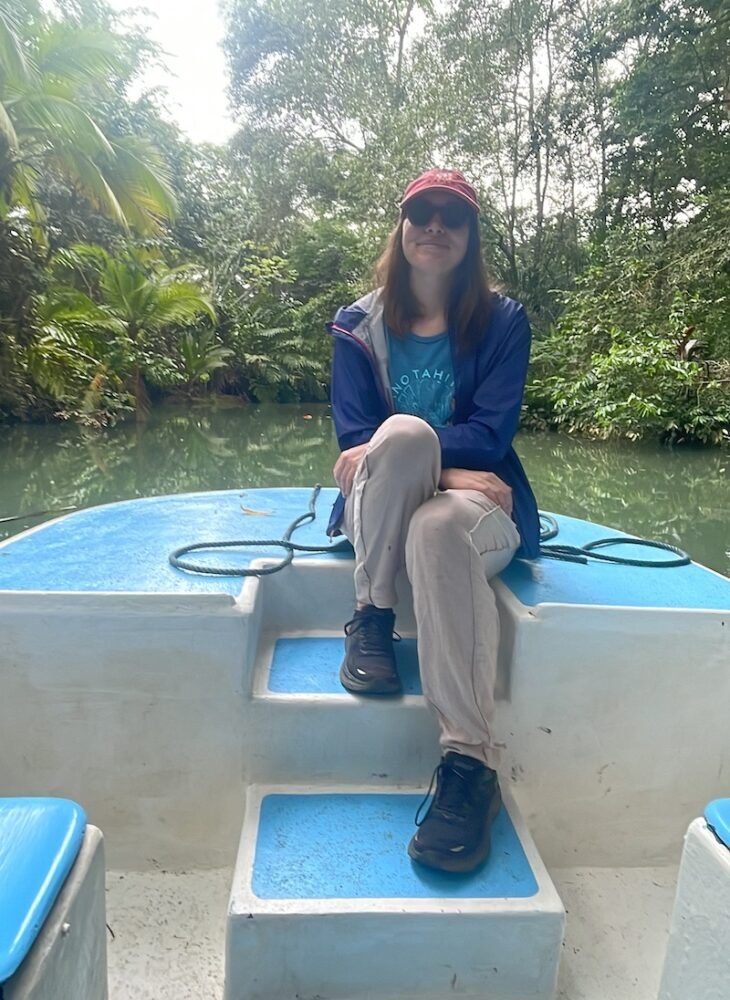 Allison Green sitting on a blue boat, with a jacket on, long pants, black sneakers, and a red hat