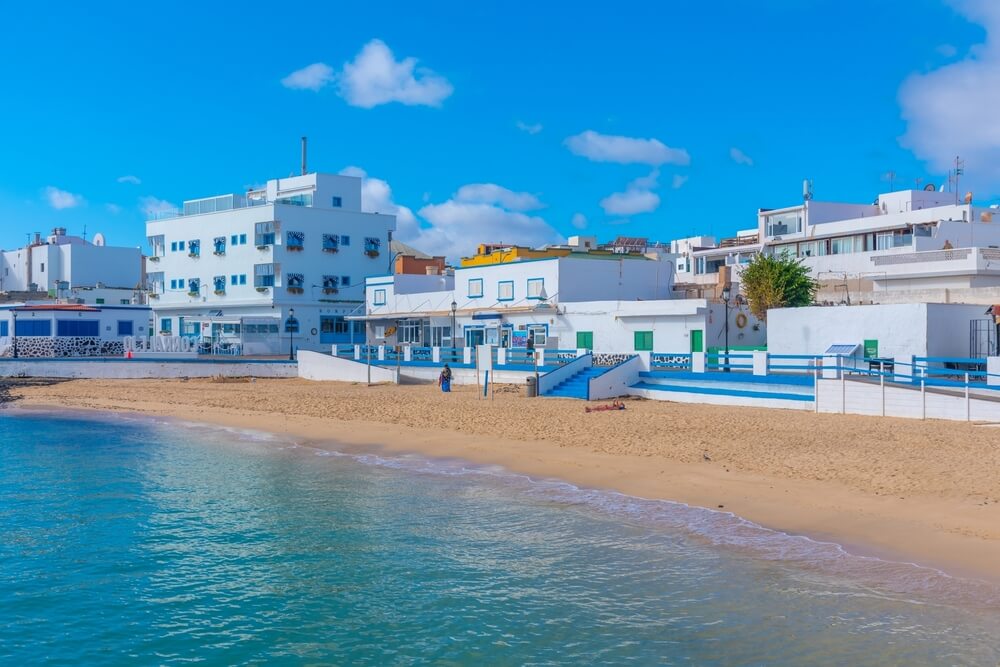 Sunny day at a beach in Corralejo, a touristic town in Fuerteventura, Canary islands, Spain. White buildings with blue detail on an empty, sandy beach with blue waters on a sunny day.