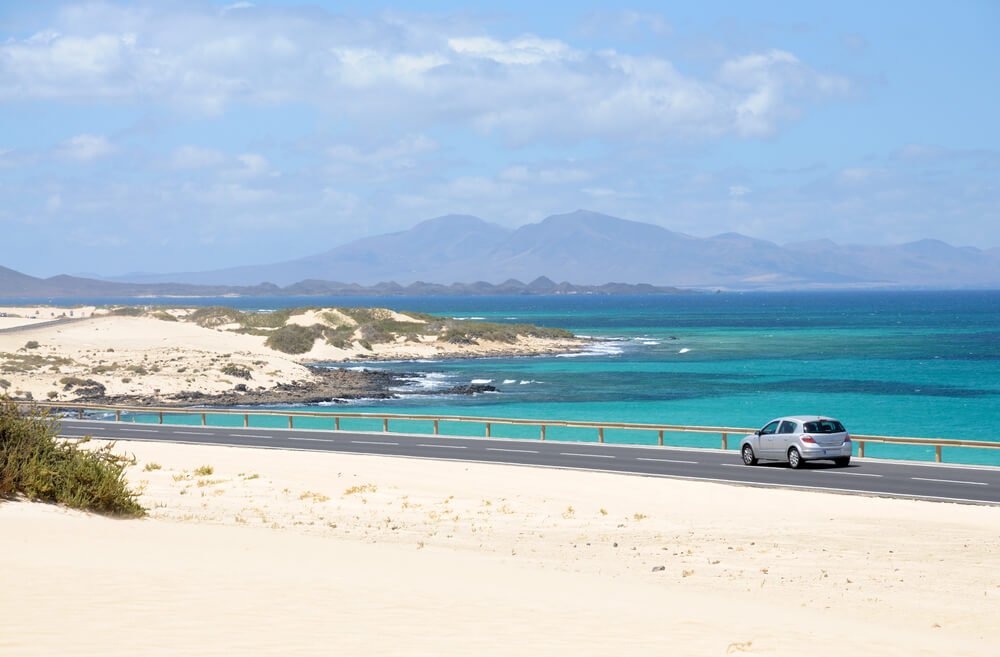 Car on the road on the paved coastal road near Corralejo in Fuerteventura, Spain, with beautiful blue waters around the sandy shores
