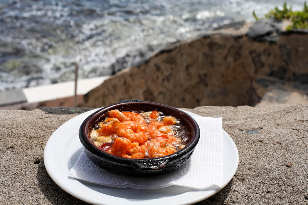 Gambas al ajillo on a plate with a napkin on the edge of a table with clifffs and beach in the view. A plate of shrimps with garlic in Fuerteventura, Canary Islands

