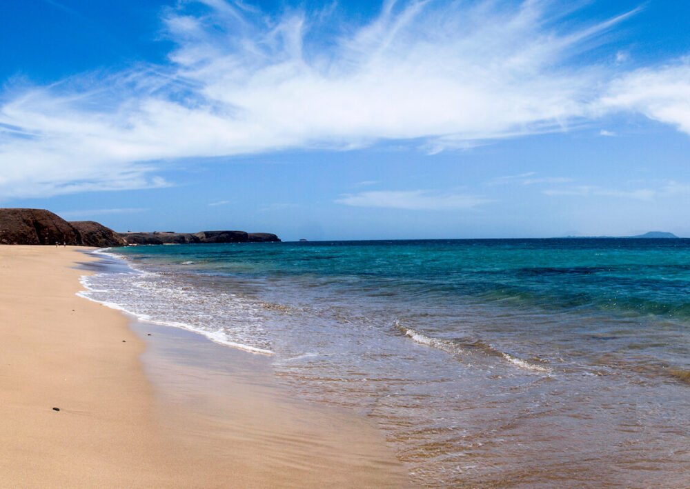 the beach of playa del pozo with golden sands, blue water, and an island visible far off in the distant horizon, on a sunny day in fuerteventura in the sand dunes