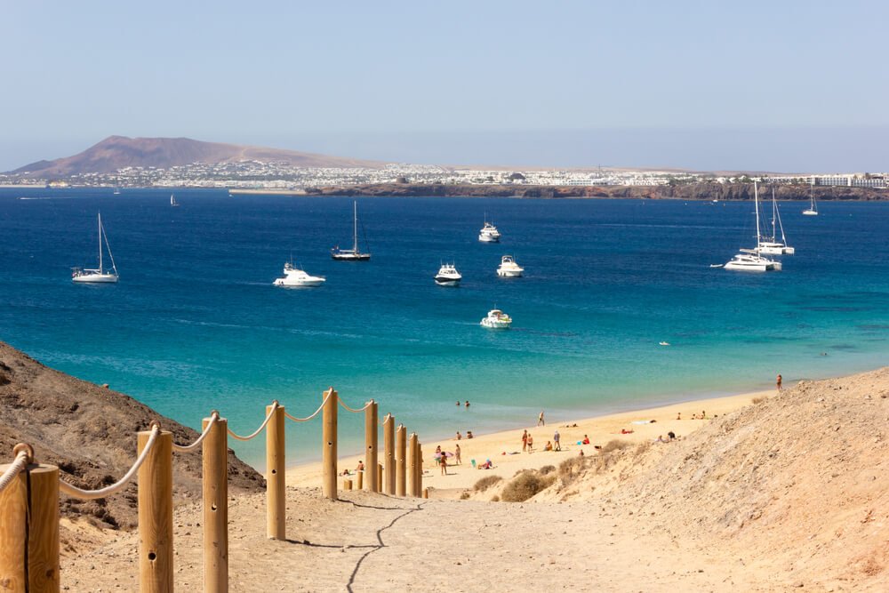 Wooden sticks fence line leading to exotic Papagayo beach in Lanzarote on sunny day. Dirt path with people on secluded bay on background by turquoise water