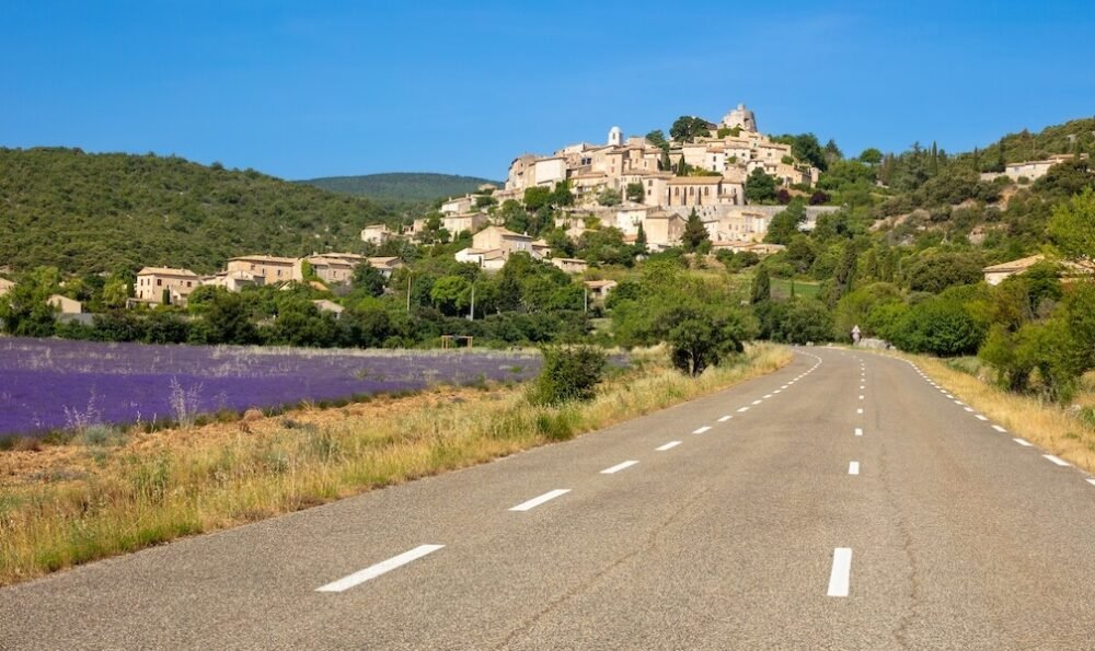 a view on the road in provence with lavender on one side of the road and a town up ahead in the distance