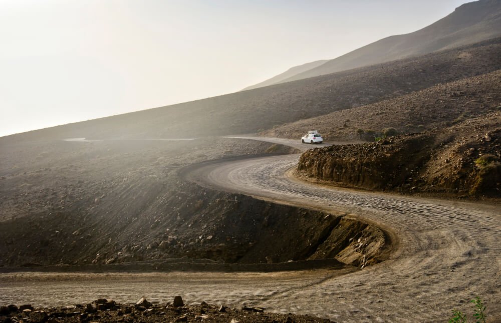 Deserted landscape with ground road on Jandia peninsula on Fuerteventura island in Spain. A white car rides along the road to the beach with surfboards on the roof. Canary Islands.
