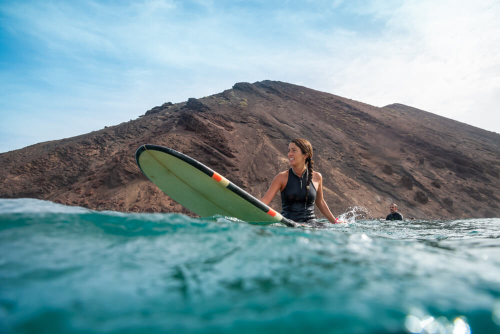Beautiful brunette woman surfing in fuerteventura, view from water level, with surfboard visible, wearing a short sleeve rashguard
