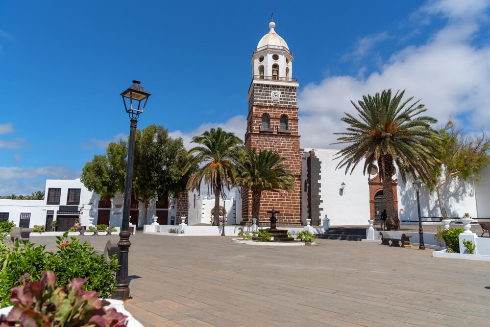 Costa Teguise village square with a church and a brick tower surrounded by streetlights Palm trees and little White Houses on a Sunny summer day. 
