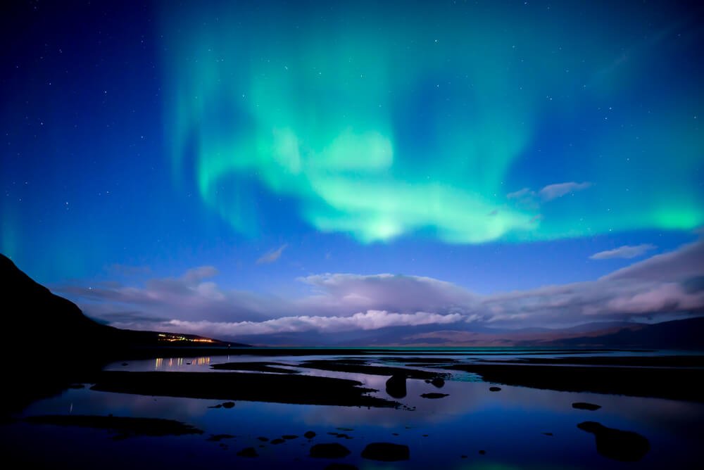 The beautiful aurora in the sky over Abisko, Sweden, with water in the foreground, a low-lying cloud in the distance, and city lights in the far distance.