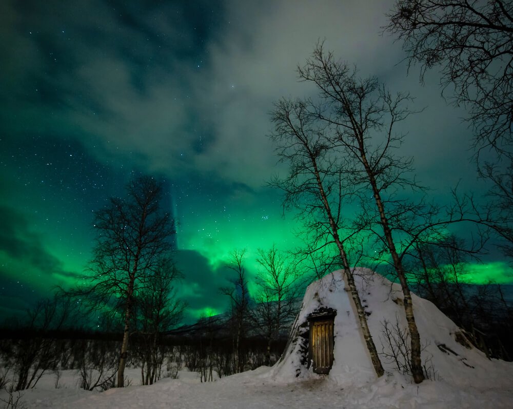 A view of the Northern lights in ABisko, with a snow-covered small house like a sauna or shed, leaf-less trees, and green sky and some clouds.