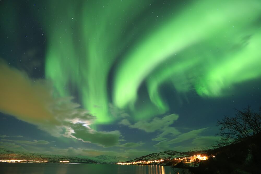 Beautiful colors of the aurora in shades of bright green, with the moon behind a cloud, streaks of cloud, and a town along the water.