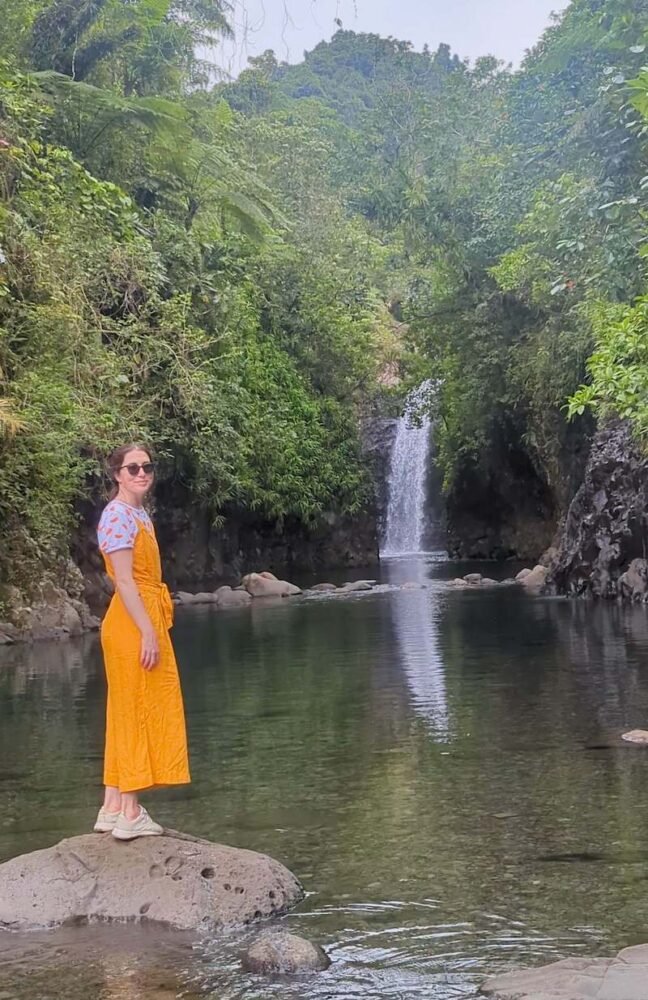 Allison Green standing in a yellow jumpsuit at the end of the Lavena Coastal Walk, where you can see Wainabau falls and two different natural pools