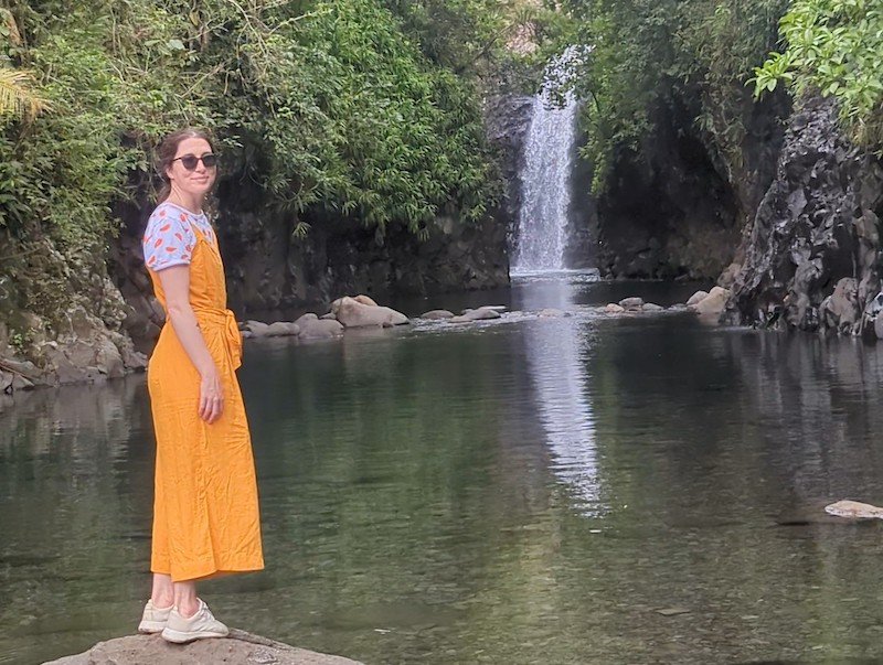 allison in front of the waterfall at the end of the lavena coastal walk, called wainabau falls, wearing a yellow romper and white shirt and shoes.