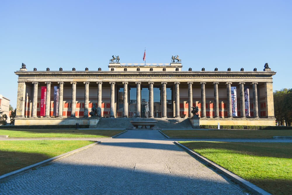 Colonnade and pillar exterior of a large museum building in Berlin with extensive grassy fields in front of it and lots of shadows, taken in mid afternoon.