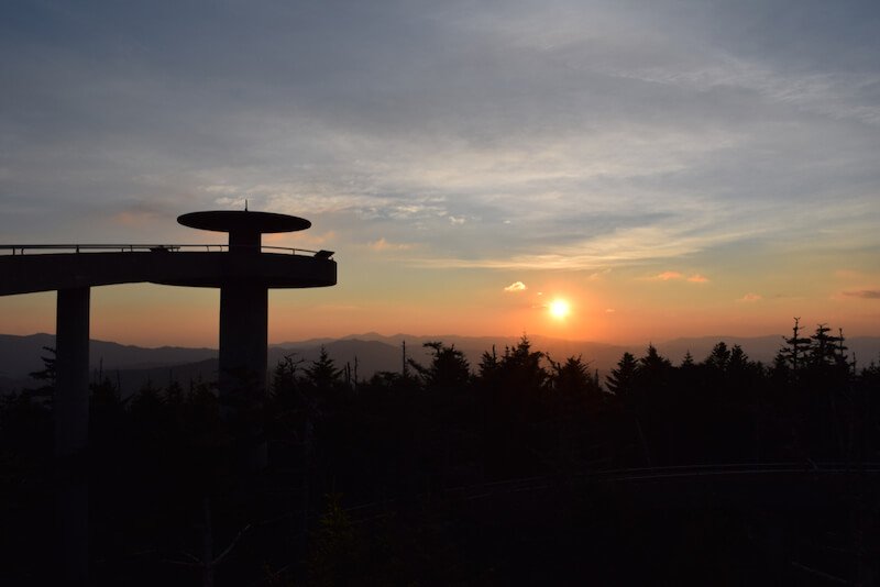 Viewpoint at Clingman Dome with a view of the sun on the horizon and pale colors lighting up the sky silhouetting against the forest and mountains.