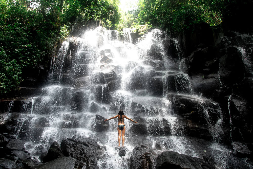 woman wearing a black bikini in front of a roaring waterfall called kanto lampo near ubud, indonesia