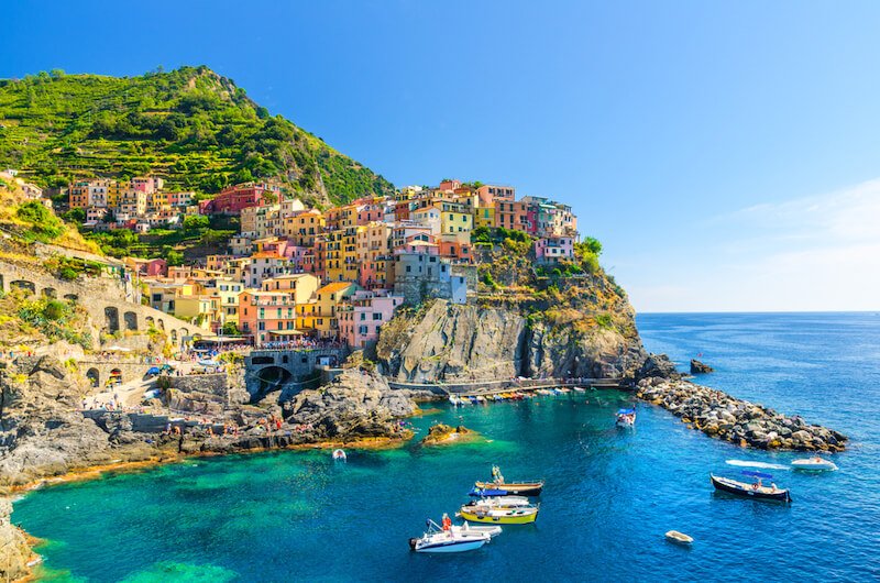 The typical Italian village of Manarola with its colorful buildings houses built on a rocky cliff, with fishing boats in the crystal clear blue waters with rocky coastline