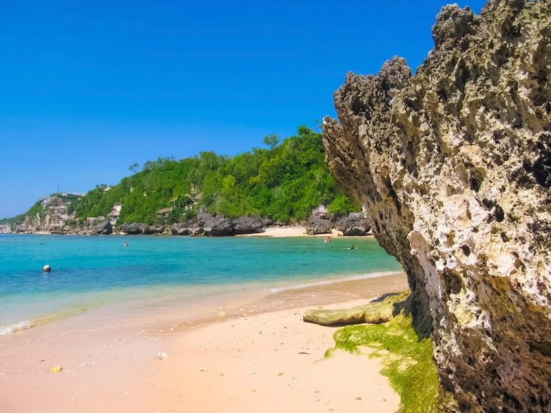 white sanad beach with turquoise and blue waters, a few people swimming in the waters and rocky cliffs and rock formations on the beach for visual interest on a sunny day on an uluwatu itinerary