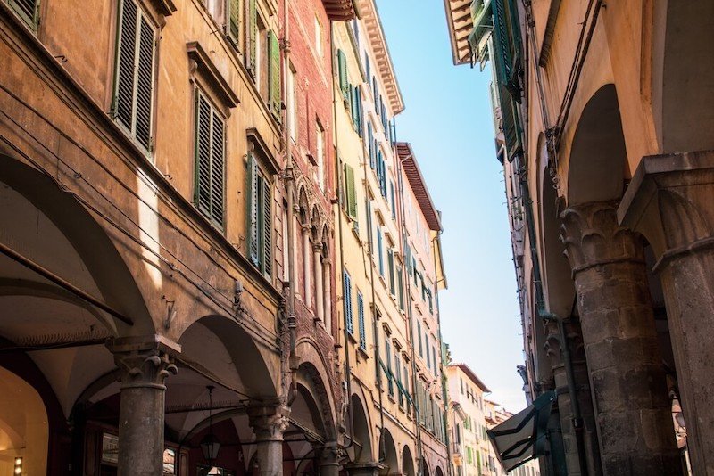 Some colored buildings on a narrow small street in Pisa, with storefronts, arches, and residential buildings up top.