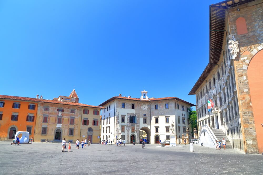 a clocktower with an arch, other brick buildings with italian flag, people walking around, orange and pastel colored facades in a piazza in pisa