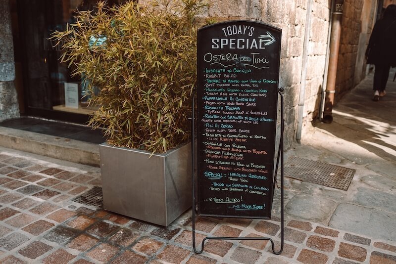  A black chalkboard menu with a list of dishes in front of a restaurant in pisa city center.