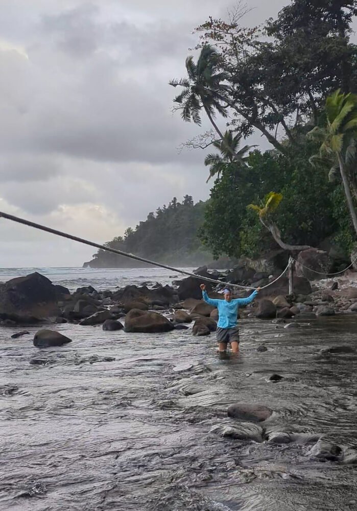 woman in a blue shirt and rolled up pants crossing the stream with her hand on a rope for balance with the ocean behind her
