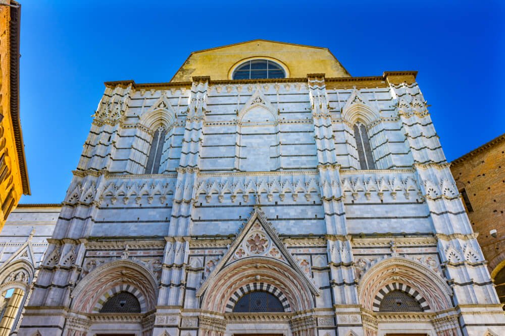 Facade of the Bapistry Cathedral in Siena, with ornate white striped marble and gold roof with circular window