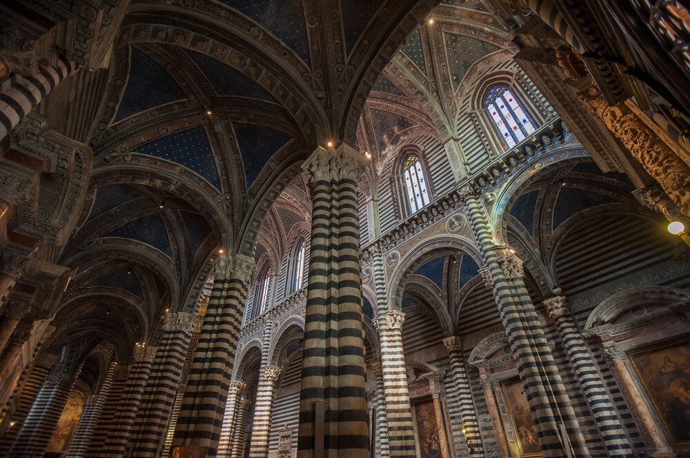 The striped interior of the Siena cathedral with a starry ceiling and stained glass and arches and domes and beautiful inlays