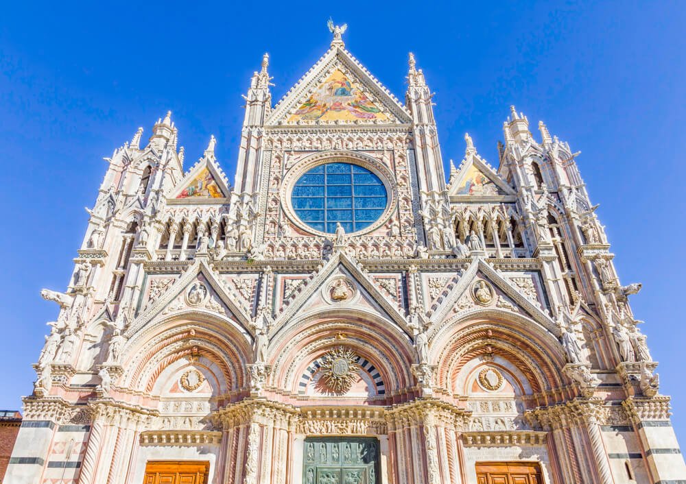 The facade of the Santa Maria Cathedral in Siena, the Duomo by other name. There are triangular pieces with paintings and lots of spires and marblework.