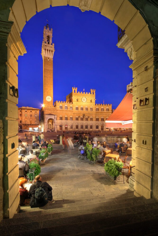 Lit up view of Palazzo Publica and Piazza del Campo through an arch with a midnight blue sky in Siena at night