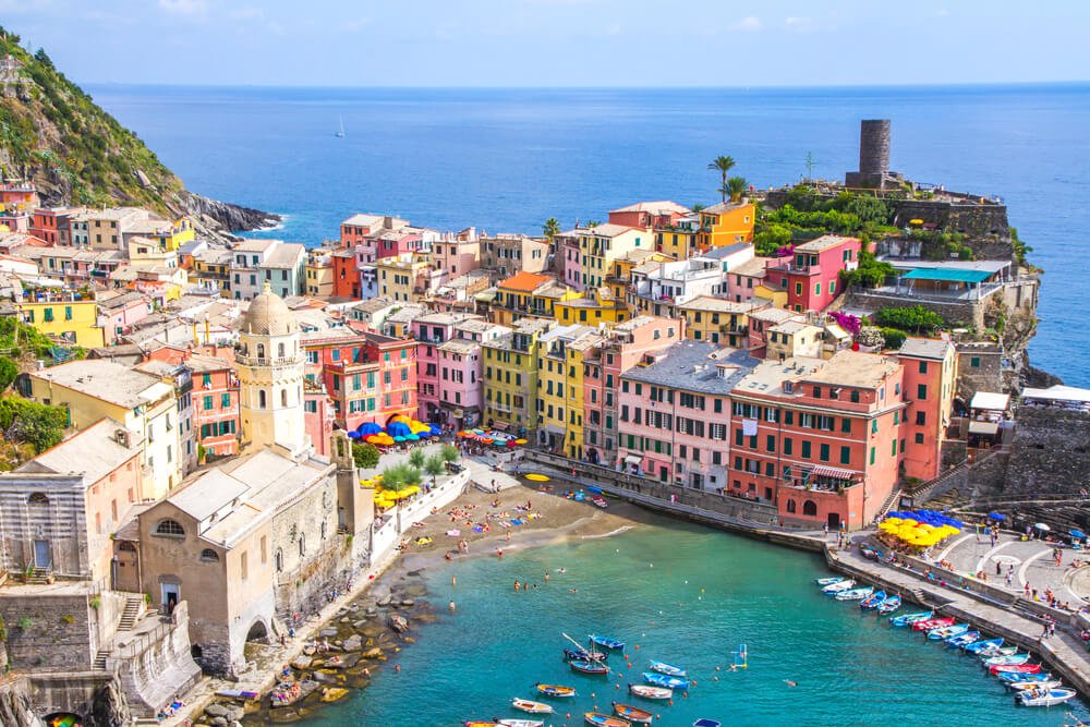 A view of the colorful coastal village of Vernazza in the Cinque Terre, with a blue sea and boat harbor, small beach, promenade with colorful houses and a tower on the hillside.
