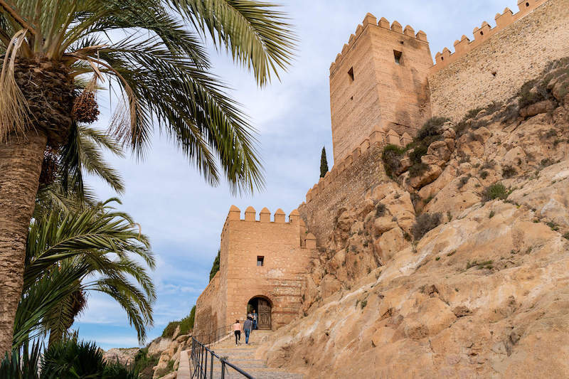 a castle view of people walking up a hill in almeria spain with palm trees in the foregorund on a day in january