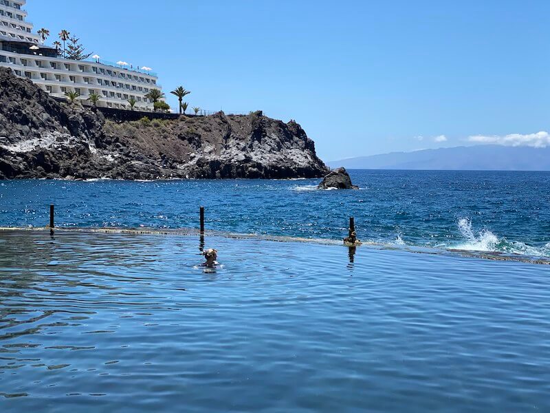 View of a natural pool in Tenerife with a cliff and ocean behind it and a hotel on the cliff side