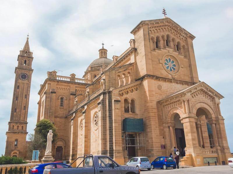 A cathedral in one of the large towns of Malta in the winter in January with a few cars out in front of the church on a partly overcast day