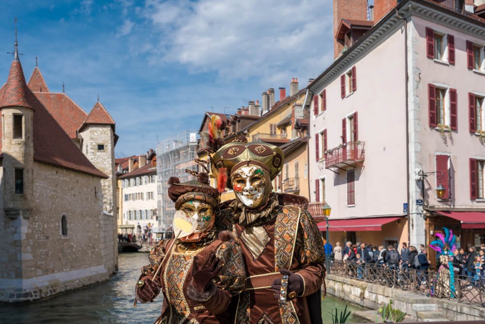 A picturesque European town scene with historic buildings and a canal. In the foreground, two individuals are dressed in ornate Venetian-style carnival costumes with intricate masks and headdresses, showcasing golden hues and detailed patterns.