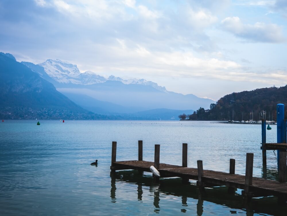 A serene winter lakeside view with a wooden dock in the foreground. A duck floats near the dock, and in the distance, snow-capped mountains give a serene winter view.  