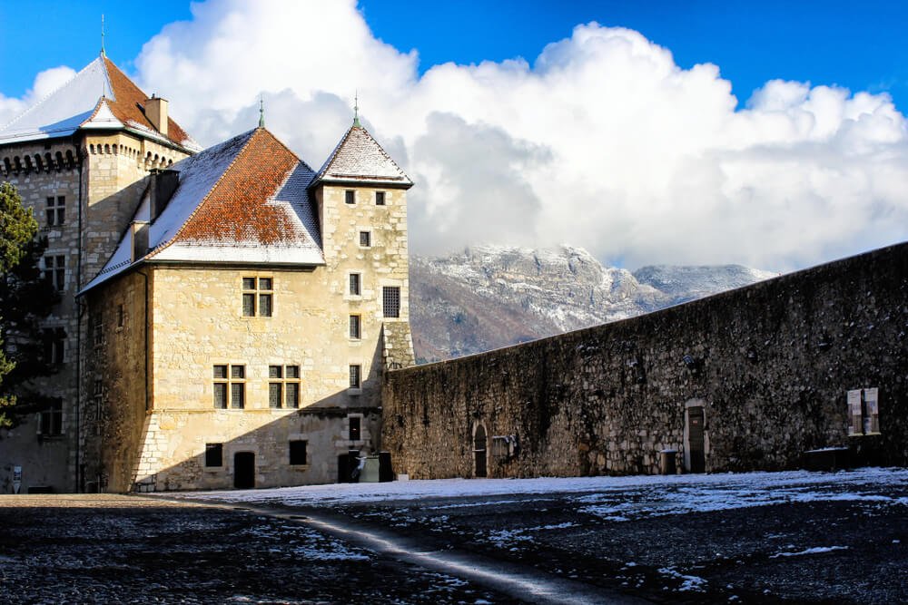 The medieval French castle in small town among snowy mountains. Snow-sprinkled square with mountains view and large, puffy clouds in winter in Annecy.