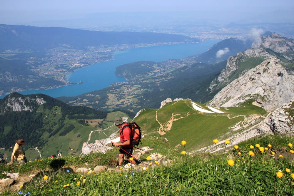 A breathtaking panoramic view from a high-altitude vantage point, showcasing a vast expanse of a blue lake below, embraced by a sprawling town and the landscape curving around its shores. Dominating the foreground are two hikers, navigating the rugged mountain terrain, adorned with patches of verdant grass and clusters of vibrant yellow flowers. The winding trails on the grassy mountain slope hint at the adventurous journey the hikers have undertaken. 
