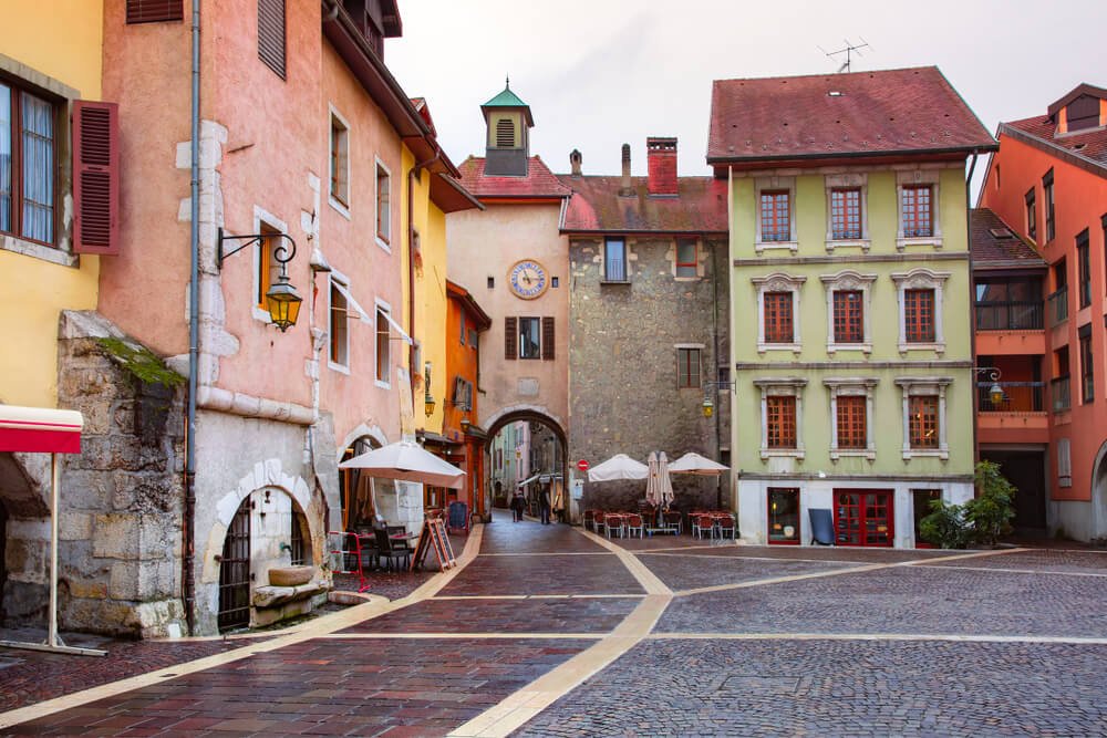 A charming European cobblestone street in Annecy, flanked by pastel-colored buildings with distinct architectural features. On the left, a vintage lantern hangs from a coral-pink building, while on the right, a lime-green structure showcases tall windows with red shutters. An archway in the distance offers a glimpse of the street beyond. The scene is peaceful, with empty cafe tables and chairs awaiting patrons.