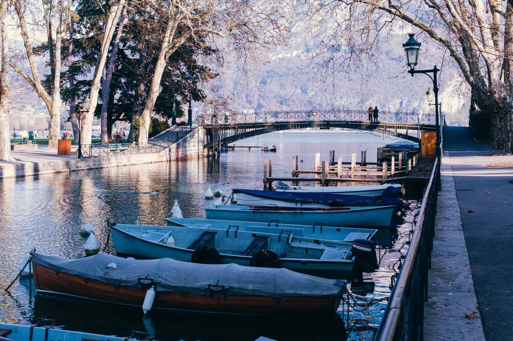 A picturesque scene of a calm canal or river, lined with tall, leafless trees. Moored boats rest along the bank, with covers protecting their interiors. In the background, an ornate bridge connects the two sides, with a couple pausing to enjoy the view in winter in Annecy.