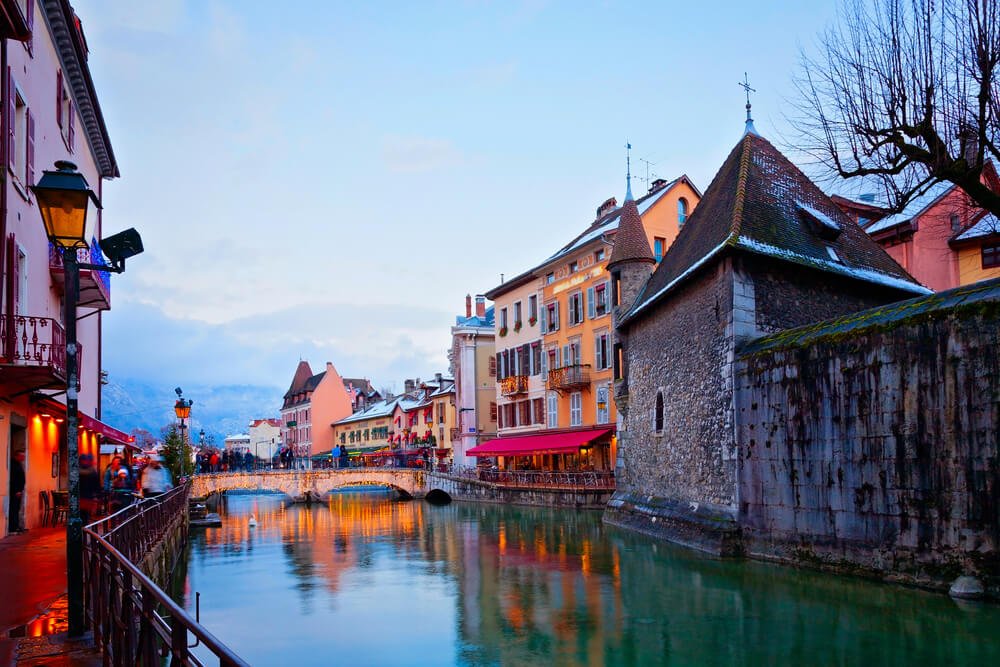 Winter landscape around dusk in Annecy with lights coming on over the canal and illuminating the old town in a festive glow.