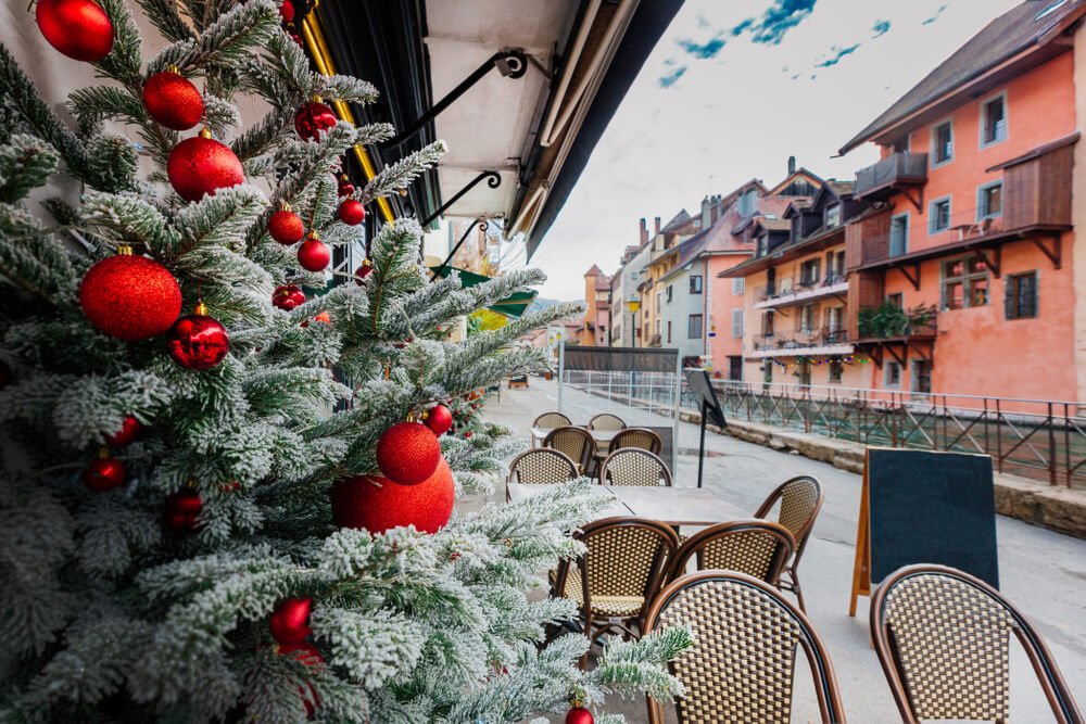 A decorated Christmas tree with red baubles stands outside a cafe with wicker chairs. Behind, colorful European buildings line a street, and a chalkboard sign is visible near the seating area, typical seating of Annecy restaurants.