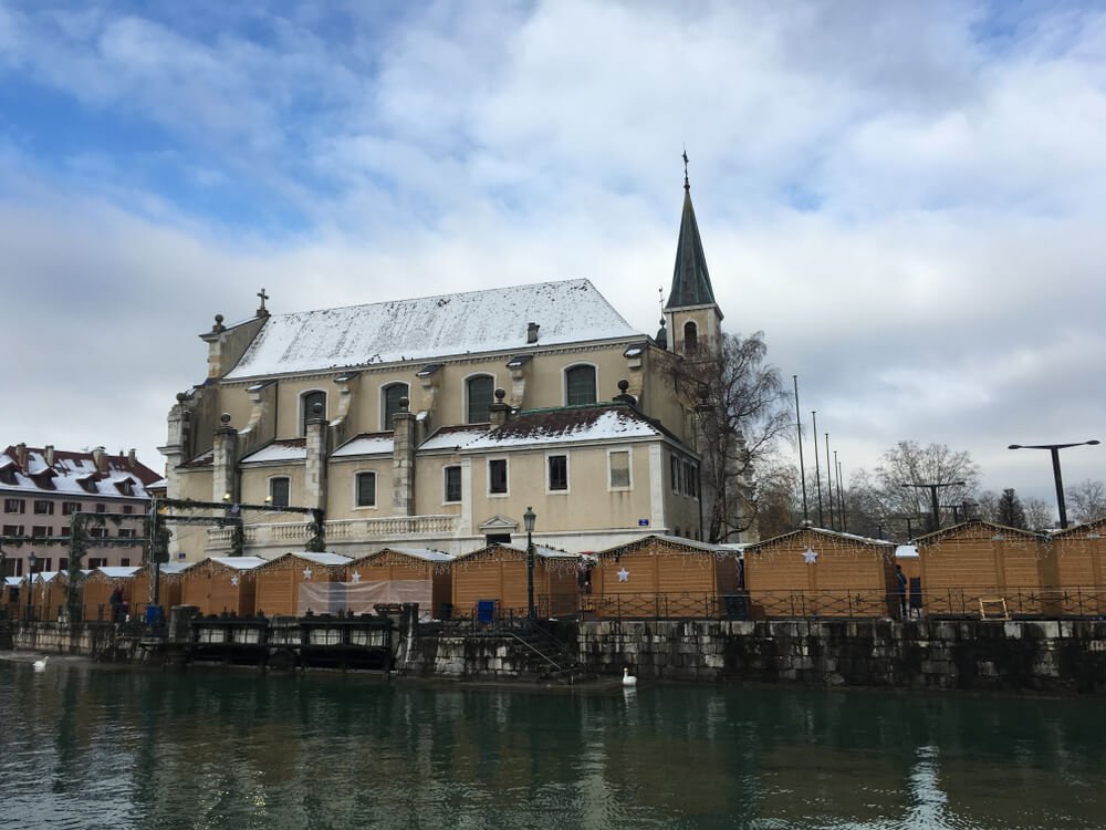 A winter scene in a European town by a canal. The focal point is a grand historic church, its façade displaying intricate architectural details and its roof lightly dusted with snow. Adjacent to the church, a slender bell tower with a green spire rises against the sky. In the foreground, the calm waters of the canal reflect the buildings, while wooden Christmas market stalls with snow-covered roofs line the embankment