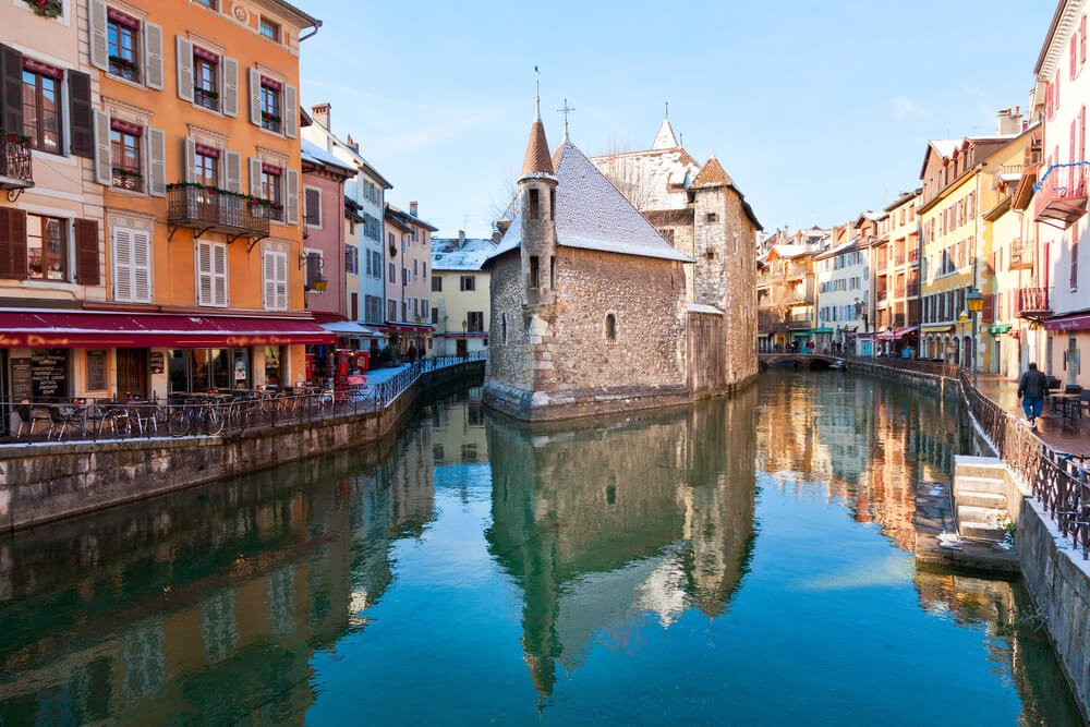 A picturesque European canal scene with colorful buildings lining the water, a stone tower, and a bridge in the background. Outdoor cafes and bicycles can be seen along the canal's edge. The water reflects the surrounding structures. There is a light dusting of snow on the roofs and sidewalks, showing a winter scene.
