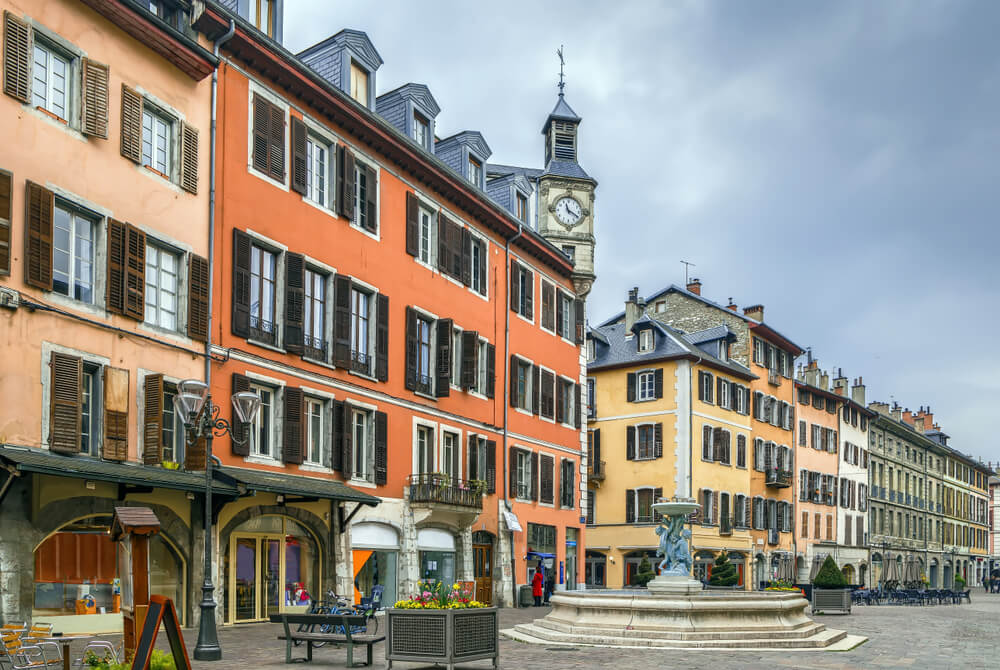 A charming European town square adorned with colorful and historical buildings. The facades of the buildings showcase a mix of warm hues like orange, pink, and yellow, with wooden shutters on windows. 