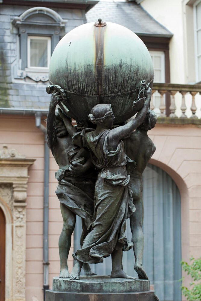 An intricate bronze statue stands prominently in the courtyard of the Bartholdi Museum. Two figures, draped in flowing robes, work in unison to support a large, tarnished sphere. The patina on the globe and figures speaks of age, adding a historical resonance to the scene. The woman on the right reaches upwards, her delicate fingers grasping a piece of the globe, while the other figure appears to be guiding or steadying it from the side. 