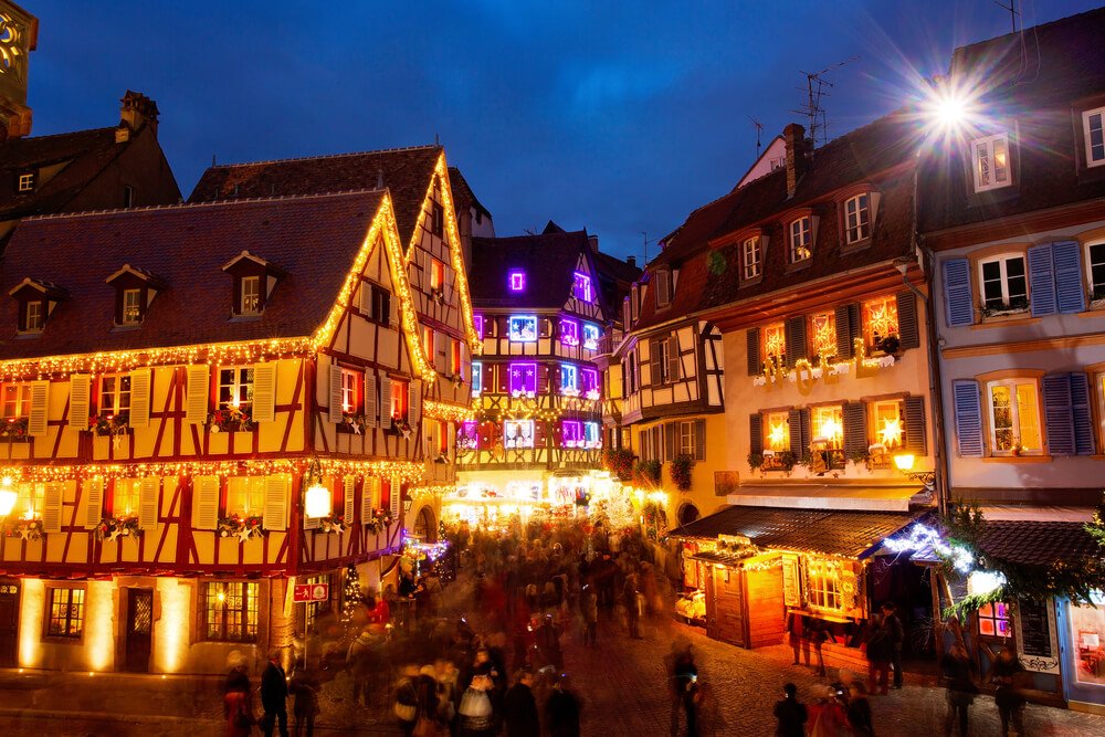 A bustling Christmas market in Colmar comes alive as night falls. Traditional half-timbered houses, adorned with twinkling fairy lights, create a warm and festive ambiance. One prominent building, illuminated in a radiant purple hue, stands out amidst the surrounding structures.