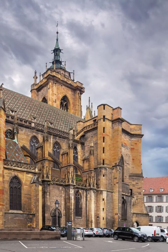 
A detailed view of a Gothic cathedral showcases its intricate stonework, ornate arches, and a prominent spire with a green finish. The structure's historical significance is highlighted by its impressive facade and tall windows. Surrounding the cathedral, modern life is evident with a variety of parked cars lining the street and a lamppost.