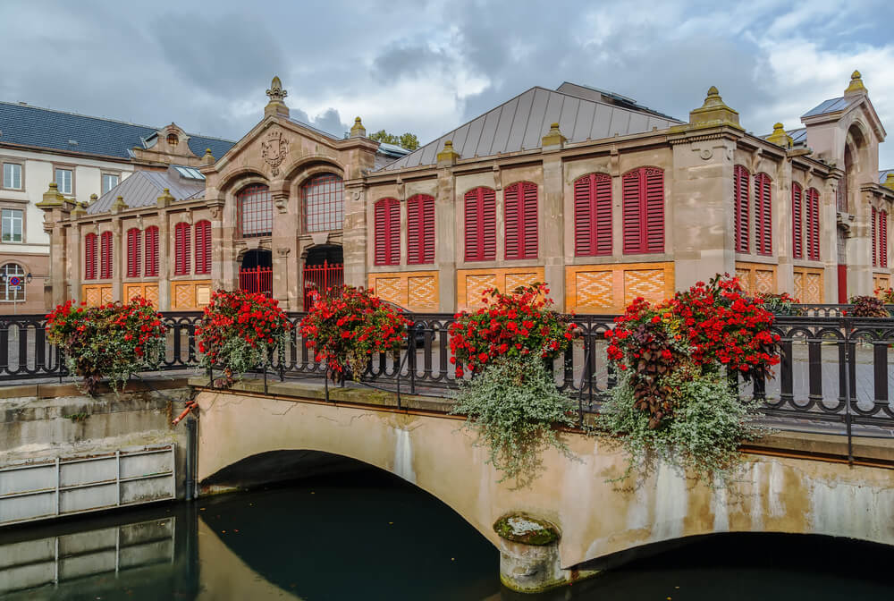 An elegant building, known as the Covered Market in Colmar, stands beside calm waters. Its beige facade is adorned with bright red shutters and large arched windows, topped by ornate stonework and decorative pediments. Overflowing flower boxes with vibrant red blooms and cascading greenery are positioned along a balustrade, adding a touch of nature to the urban setting. An arching stone bridge spans the waterway in the foreground, reflecting the city's blend of architectural charm and functional design.