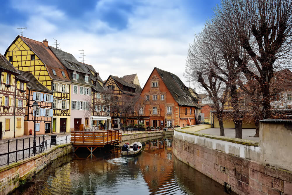 A boat going down one of the canals of Colmar in the winter, surrounded by leafless trees showing the season, yellow and pastel colored half-timbered houses.