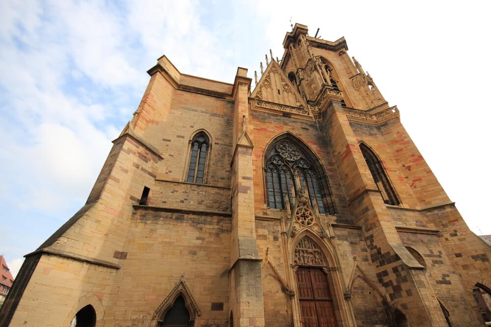 The facade of the orange/sand-colored Dominican church of colmar on a partly cloudy day.