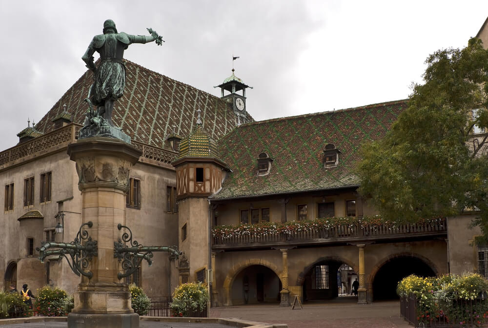 A grand historical building, known as the Koïfhus, stands majestically in the heart of Colmar. Its distinct green and red patterned tile roof captures immediate attention. A central bronze statue, mounted atop a sturdy stone pedestal, dominates the courtyard. The figure, adorned in period attire, extends an arm forward.